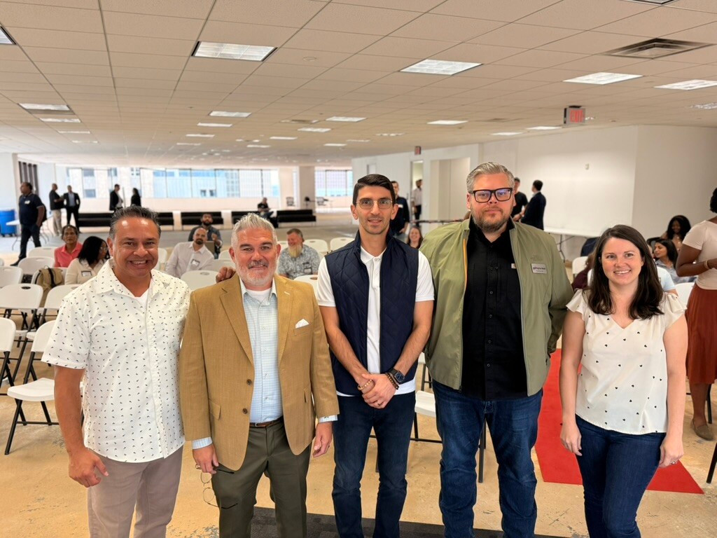 Five attendees pose together in the event space, with rows of white chairs and other attendees behind them.